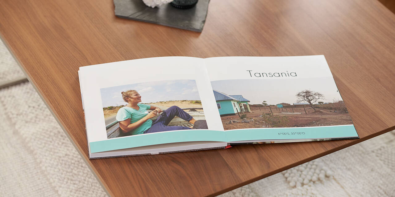 Auf einem Holztisch liegt ein aufgeschlagenes Fotobuch mit Bildern und der Überschrift "Tansania". Über dem Fotobuch steht ein dunkles Tablett mit einer Kerze und Muschel. Unter dem Tisch ist ein heller Teppich zu sehen.